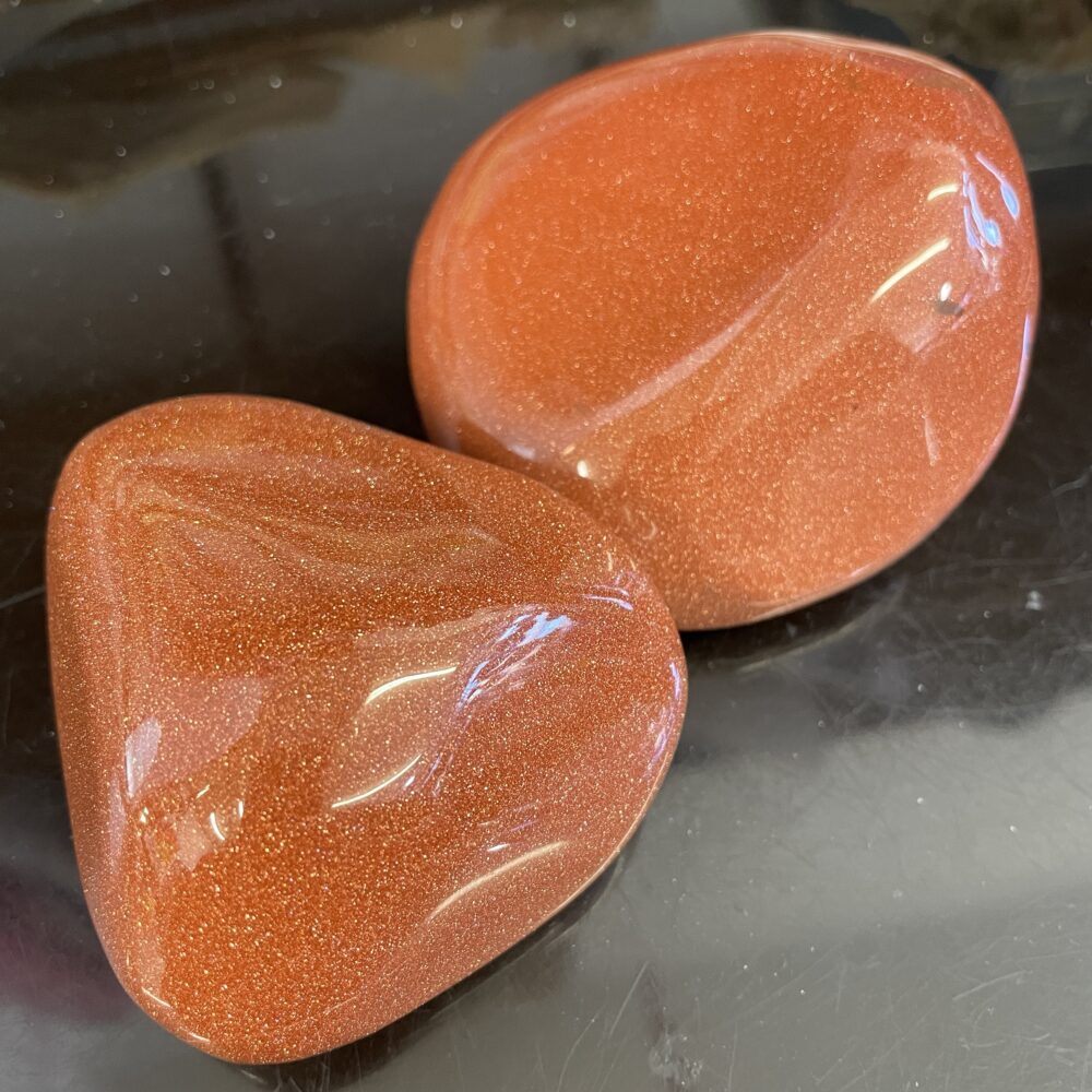 Two Goldstone Healing Therapy Stones, featuring a sparkling, glittery surface, are displayed on a reflective dark surface, making them perfect as healing therapy stones or eye-catching display specimens.