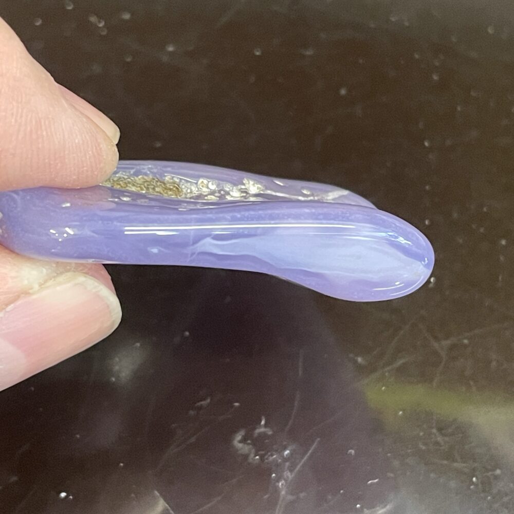A person holds a smooth, translucent, curved object with a light purple hue against a dark background. This mesmerizing stone is the Rare Holly Blue (Calapooia) Agate 31g or 155cts, captivating in its delicate beauty and rarity.