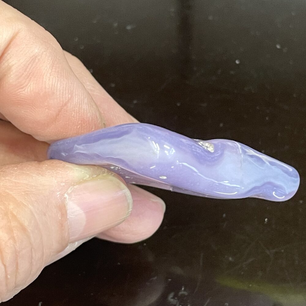 A hand holds a smooth, elongated purple stone with wavy white patterns, resembling the Rare Holly Blue (Calapooia) Agate 31g or 155cts.