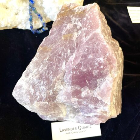 Huge boulder of Lavender Cherry type Rose Quartz, weighing 11.1 lbs, displayed on a dark surface with a label in front.