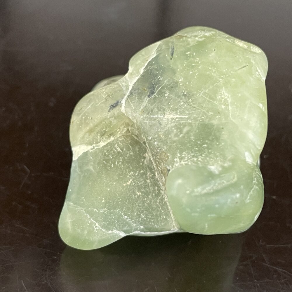 The Prehinite Natural Shaped Tumble Polished, 68.4g/2.4oz, is a smooth, light green stone with surface scratches on a dark, glossy background.