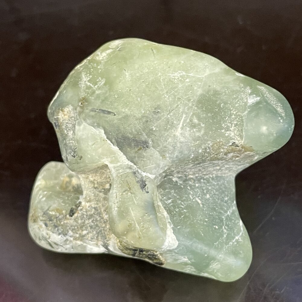 A Prehinite natural shaped tumble polished stone, weighing 68.4 grams (2.4 oz), with a translucent green hue and surface imperfections, displayed on a dark background.