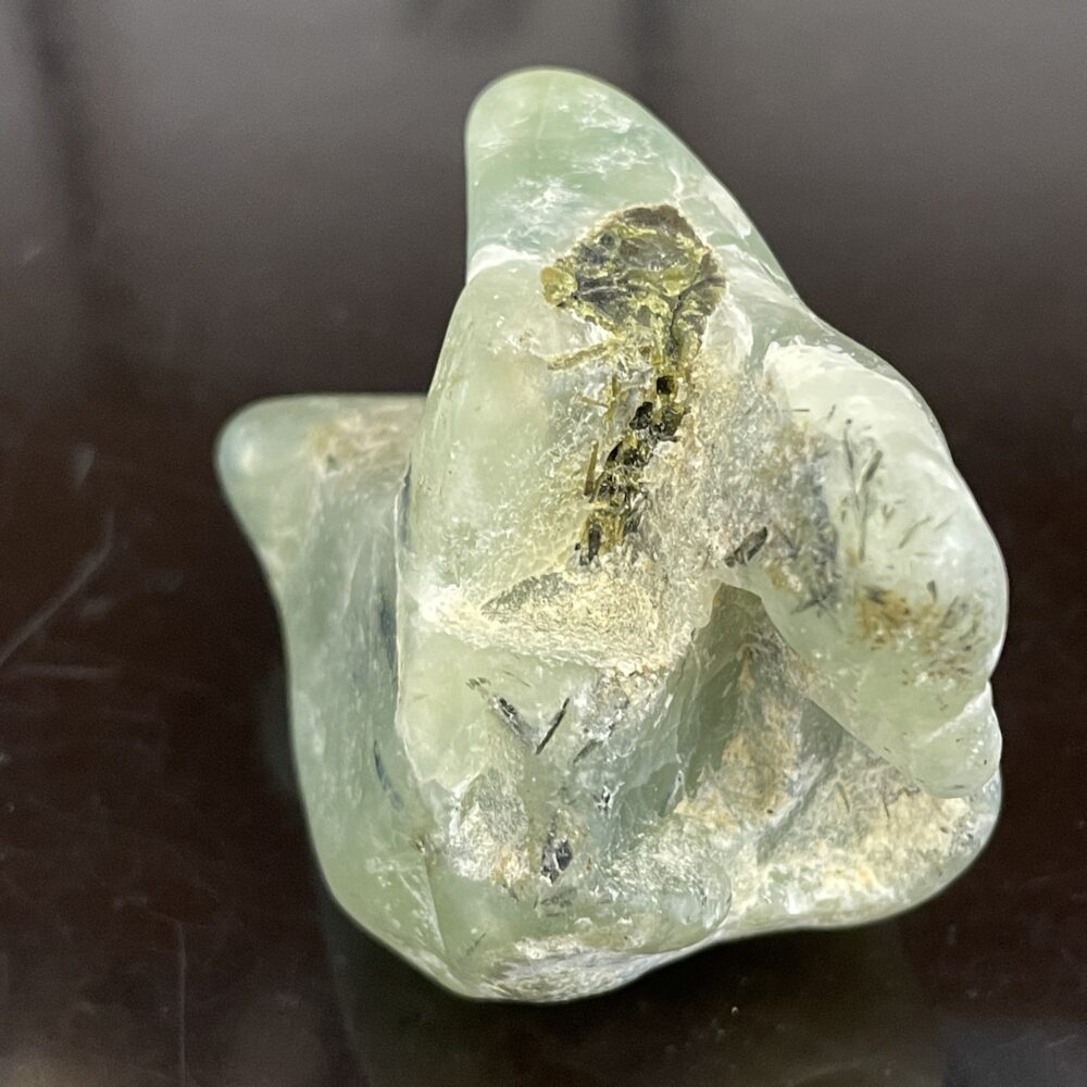 A rough, translucent green Prehinite tumbled specimen with yellowish and black inclusions, on a dark reflective surface.