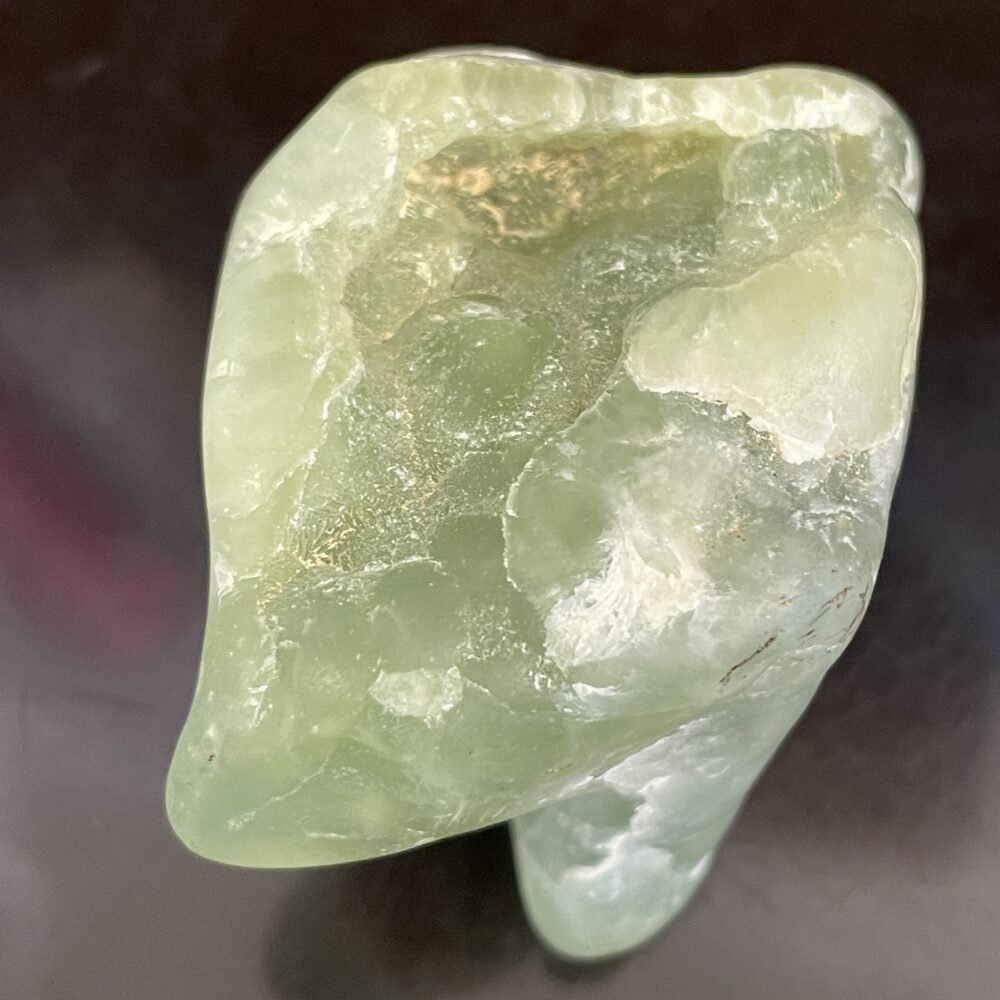 A close-up of the Prehinite Natural Shaped Tumble Polished stone, semi-translucent with a smooth finish, on a dark background.