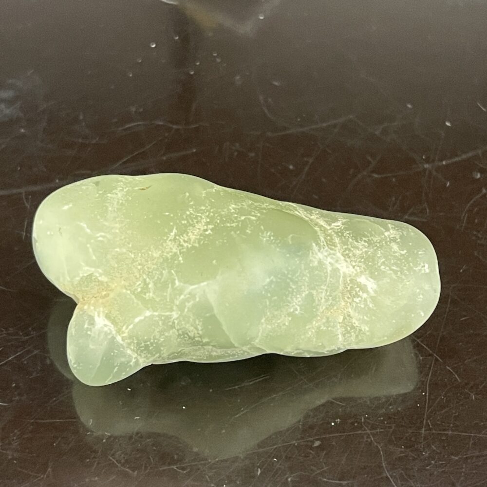 A smooth, pale green Prehinite natural shaped tumble polished stone (68.4 grams/2.4 oz) sits on a reflective dark surface.
