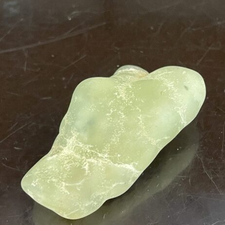 A pale green Prehinite Natural Shaped Tumble Polished sea glass fragment, 68.4g/2.4oz, rests on a dark surface with irregular edges.