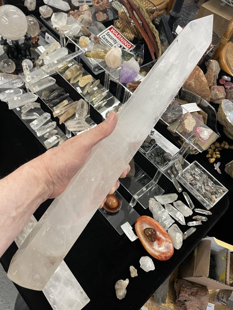 A hand holds the Rare Self Healed Lemurian Quartz Crystal Showing Hematite (5.5 long) above a table displaying assorted crystals, stones, and minerals.