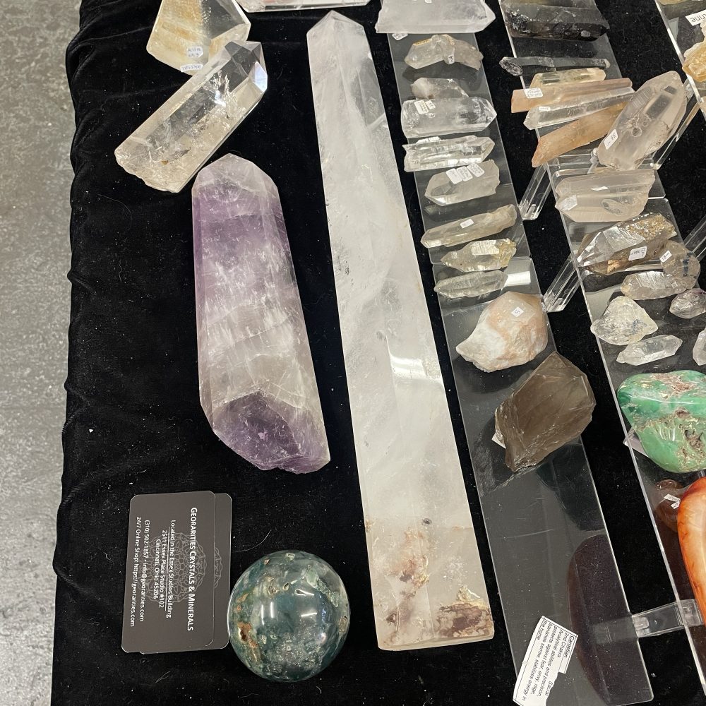 A rare self-healed Lemurian Quartz Crystal, showing hematite and measuring 5.5 inches long, is displayed among other polished and raw crystals on a black cloth at a mineral vendor’s table.