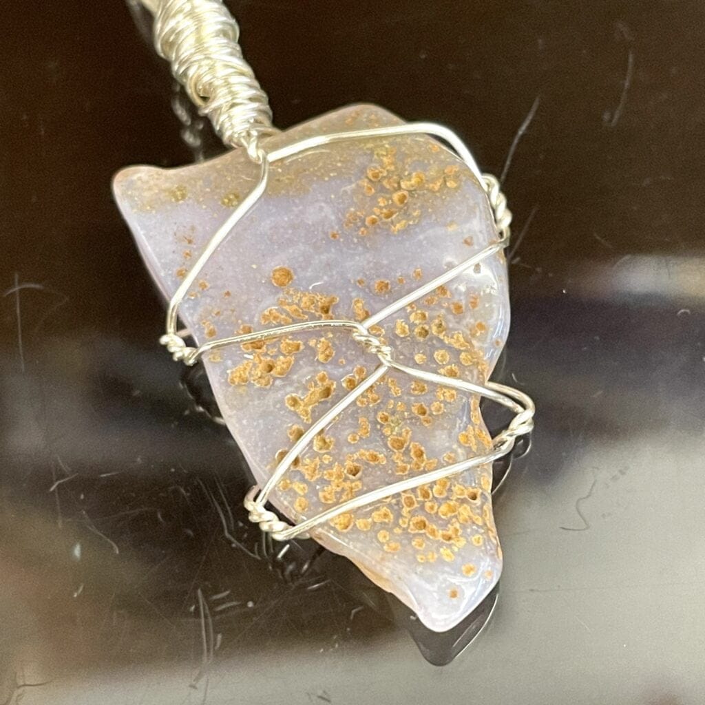 The Rare Holly Blue Agate Pendant Wire Wrap features a unique stone with yellowish-brown speckles, elegantly wire wrapped in silver and showcased on a glossy black surface.