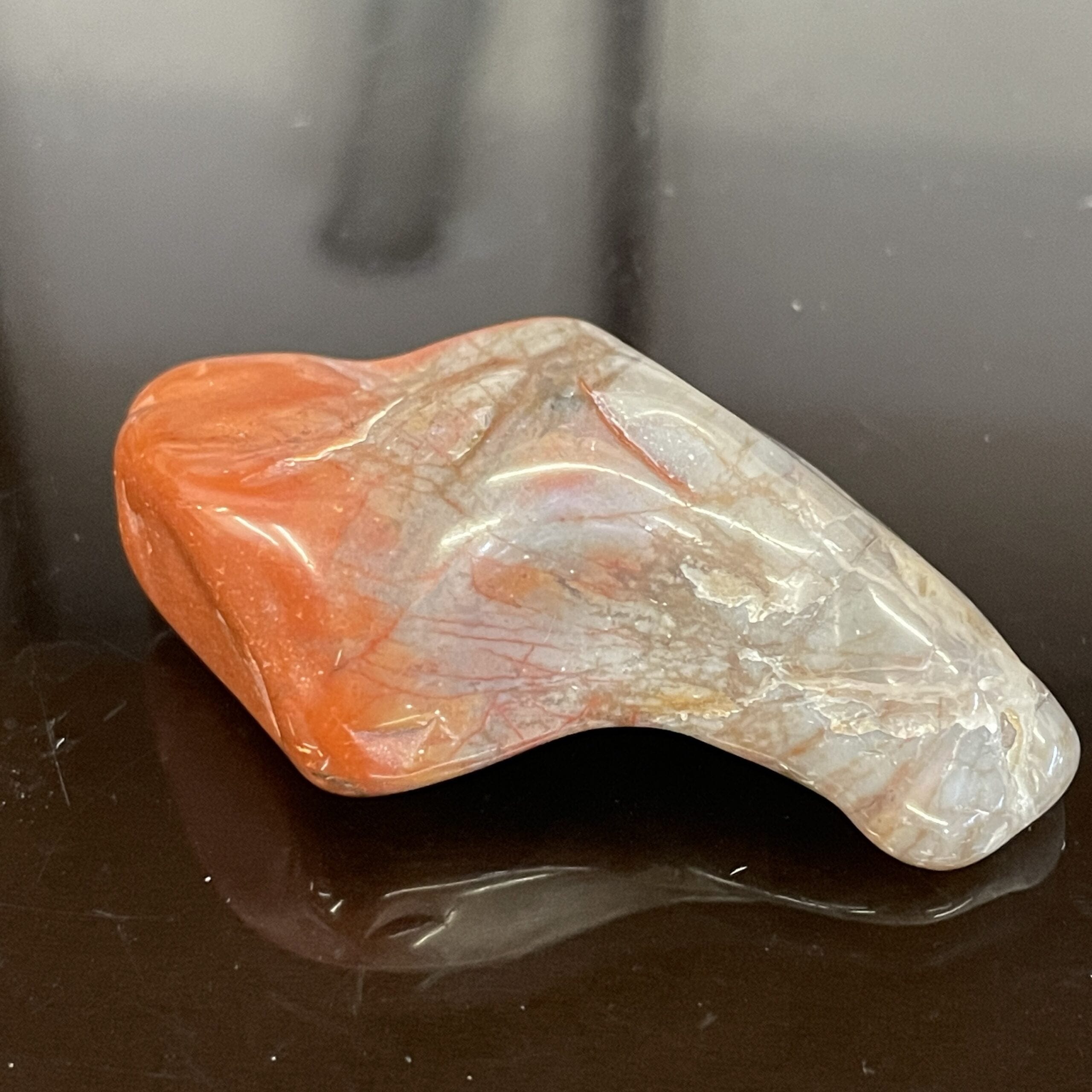 The Arizona Rainbow Petrified Wood Polished Chunk Specimen, with irregular shapes and red, orange, and white hues, rests on a glossy black surface.