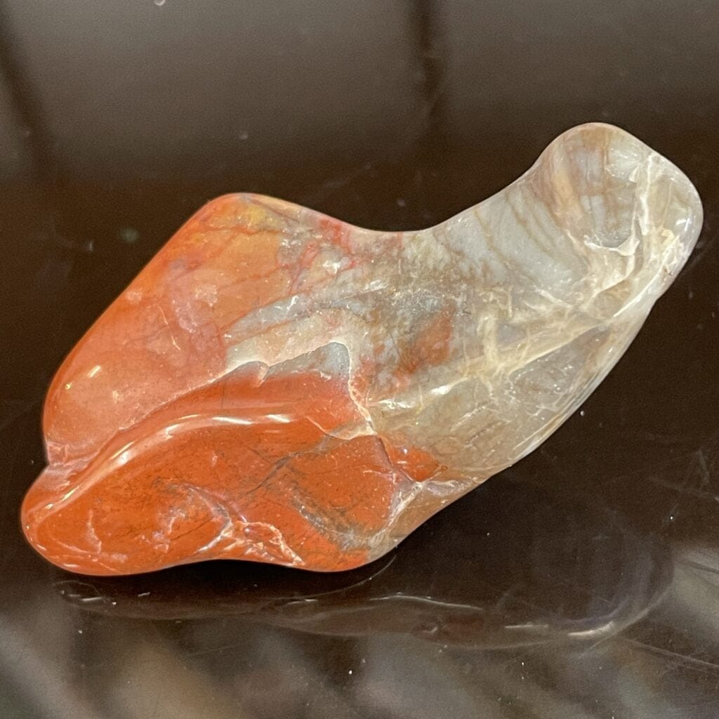 The Arizona Rainbow Petrified Wood Polished Chunk Specimen 1 oz (29g), with vibrant red, orange, and gray hues, is displayed on a reflective black surface.