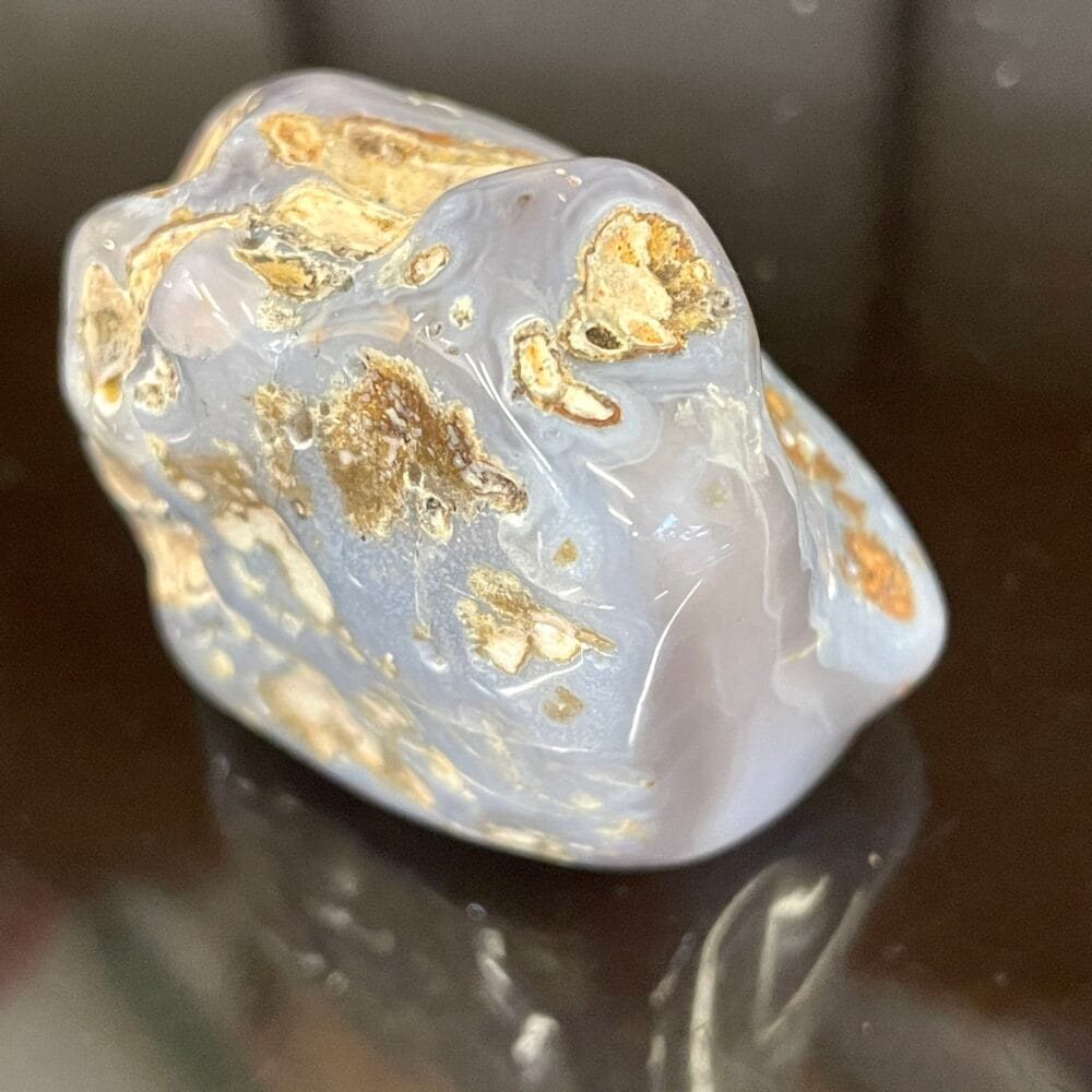 A polished Rare Holly Blue Agate 8g, known for enhancing psychic abilities and displaying smooth gray and beige patterns, rests on a dark reflective surface, exuding mystical charm.