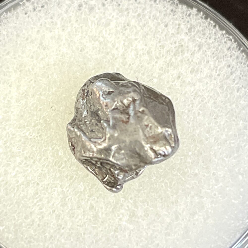 The IM23 Campo del Cielo Iron Meteorite, a 1.53g metallic specimen with an irregular shape, rests on a white textured background.