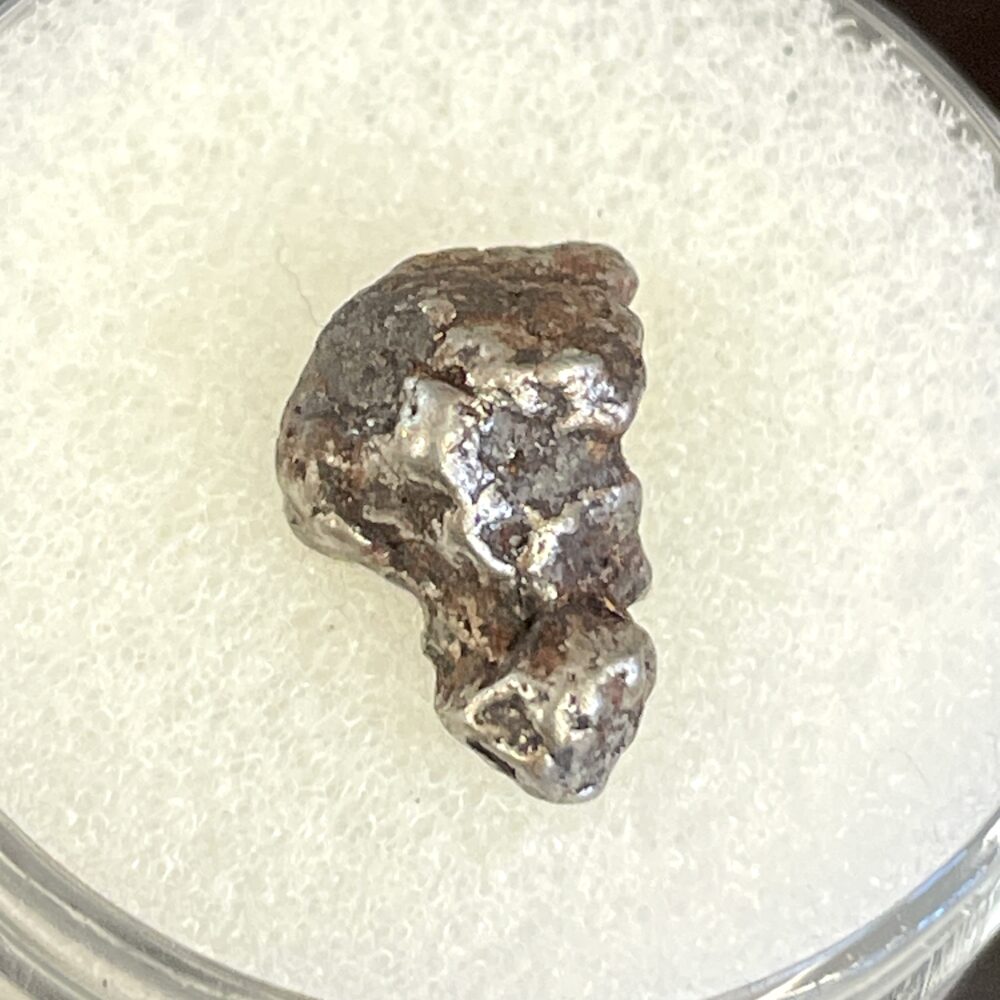 The IM26 Campo del Cielo Iron Meteorite Nice 1.55g Specimen, a small, irregularly-shaped nugget, is displayed on a white textured surface.