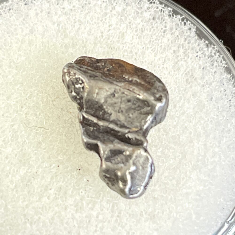 The IM26 Campo del Cielo Iron Meteorite Nice 1.55g Specimen, a small, irregularly shaped sample, rests on a white textured surface.
