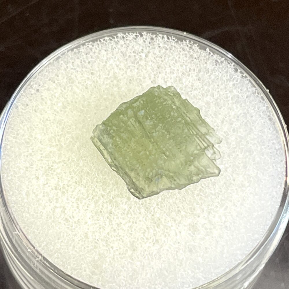 A green, translucent Moldavite crystal weighing 0.58g from Chlum, CZ is displayed on a white foam pad in a clear container.