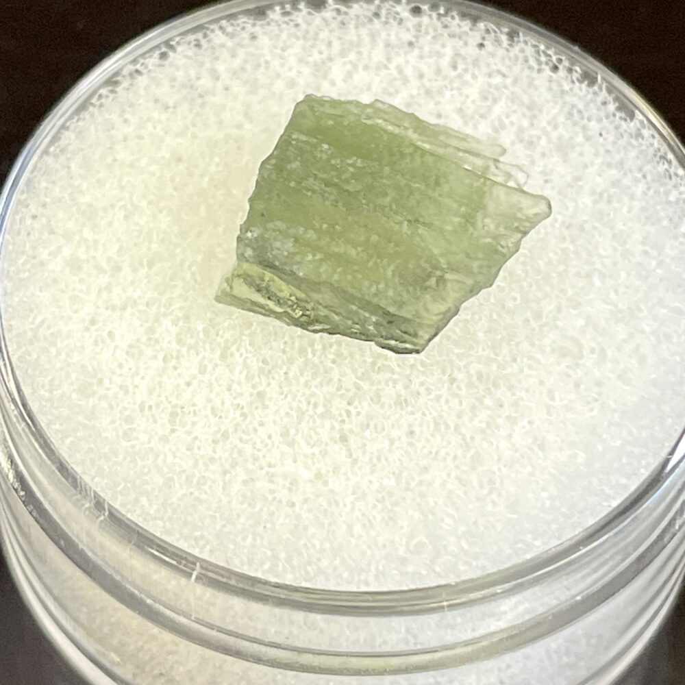 A .58g Moldavite from Chlum, CZ rests on a white textured surface inside a clear round container.