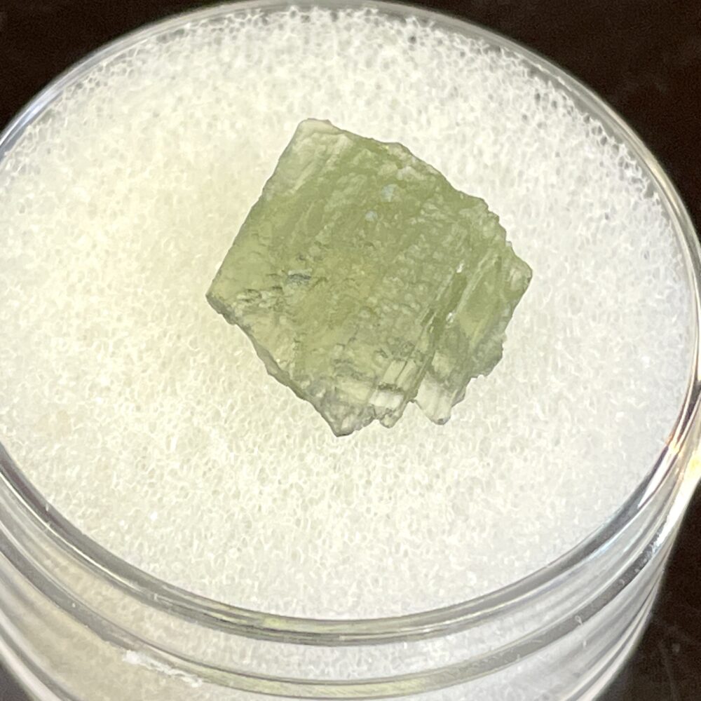 A green, translucent Moldavite from Chlum in CZ, weighing .58g, is displayed on a white foam pad inside a clear round container.