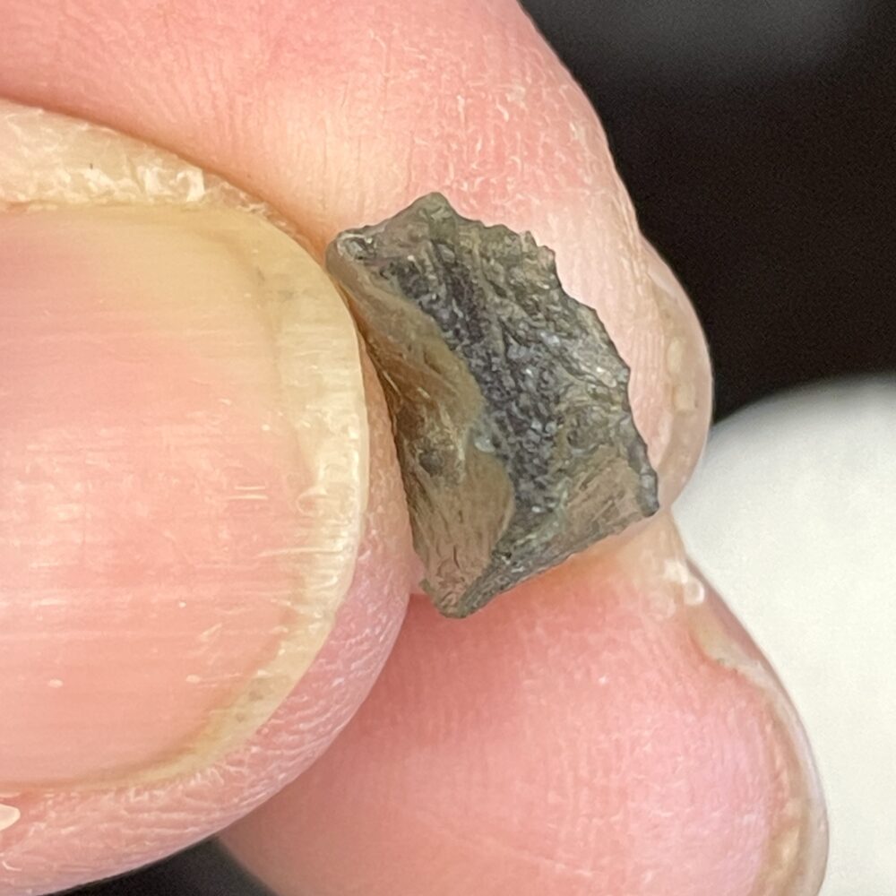 A close-up of fingers holding a small, dark object, similar in size to Moldavite from Chlum, CZ, weighing 0.58g.