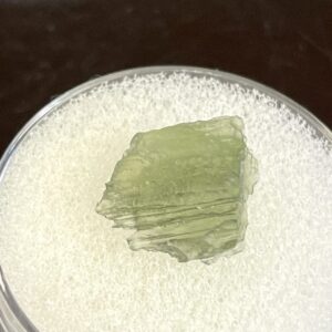 A 0.58g Moldavite from Chlum, CZ with a rough, jagged surface rests on white foam inside a clear container.