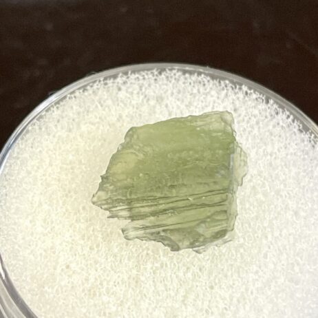A 0.58g Moldavite from Chlum, CZ with a rough, jagged surface rests on white foam inside a clear container.