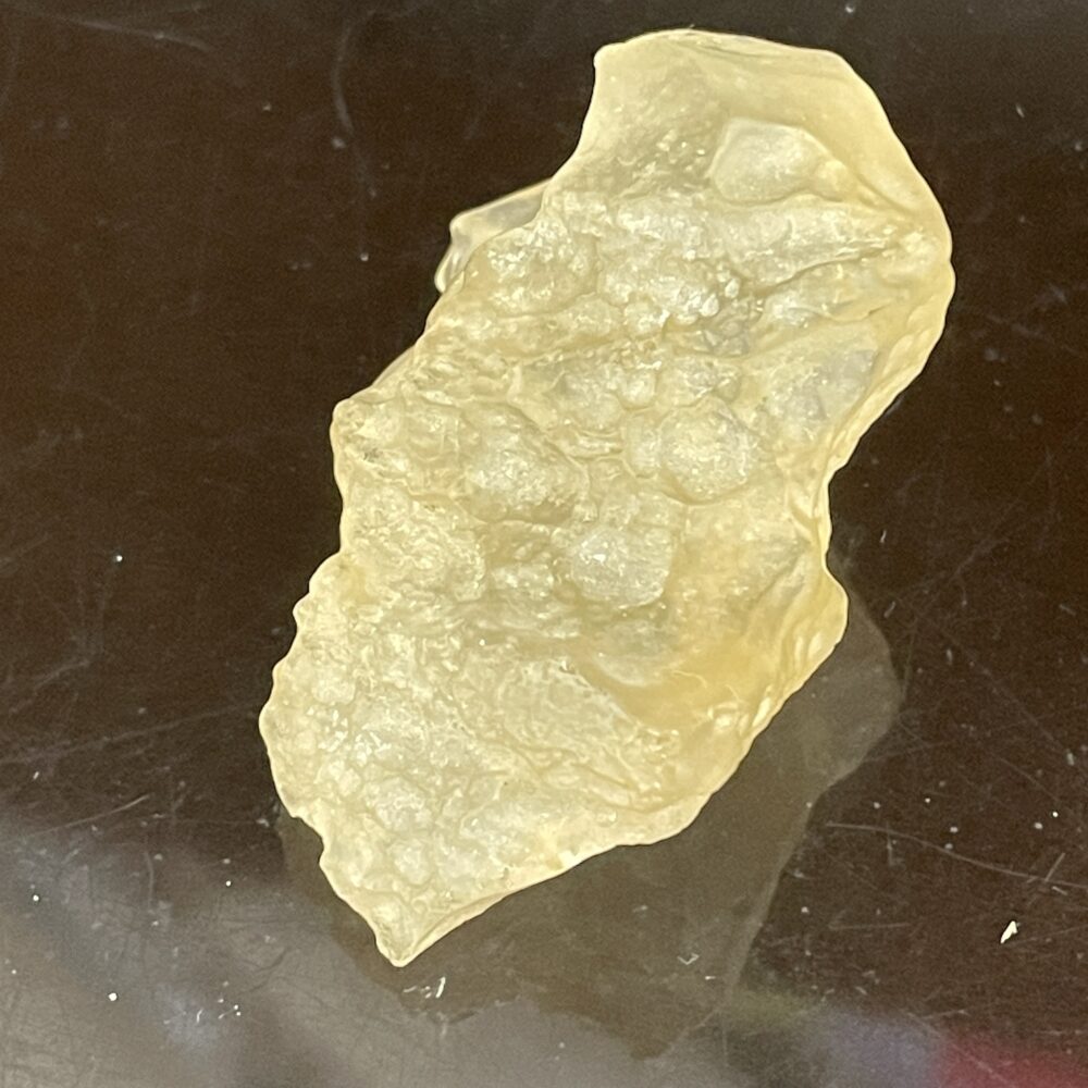 A rough, translucent, pale yellowish stone with an irregular surface, displayed on a dark reflective background.