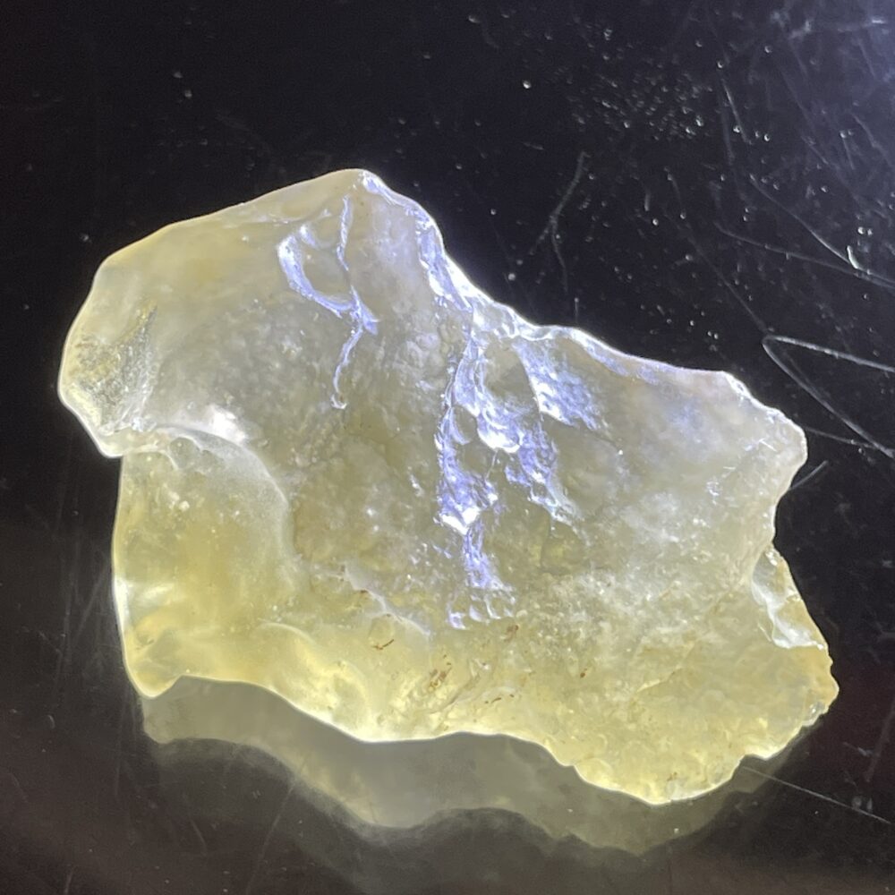 A translucent, irregularly shaped yellowish mineral or resin lies on a reflective black surface.