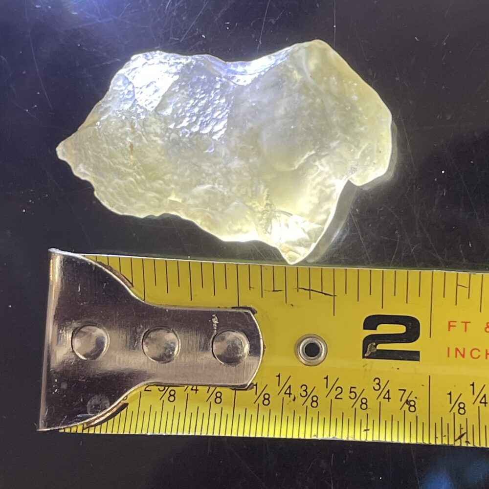 A translucent, irregularly shaped yellowish rock is placed above a yellow measuring tape, showing the rock is approximately 2 inches long.