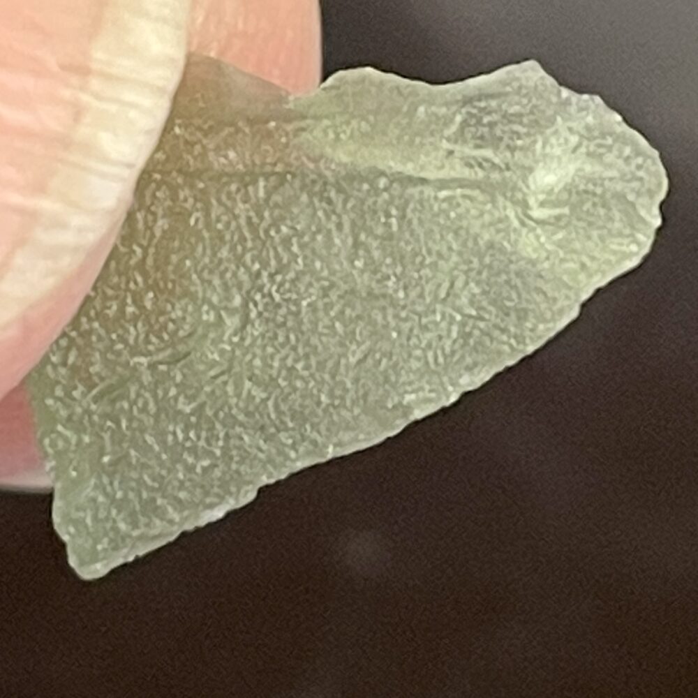 A close-up of fingers holding Moldavite From Chlum in CZ (.47g or 2.50 ct), highlighting its pale green color, irregular shape, and textured surface.