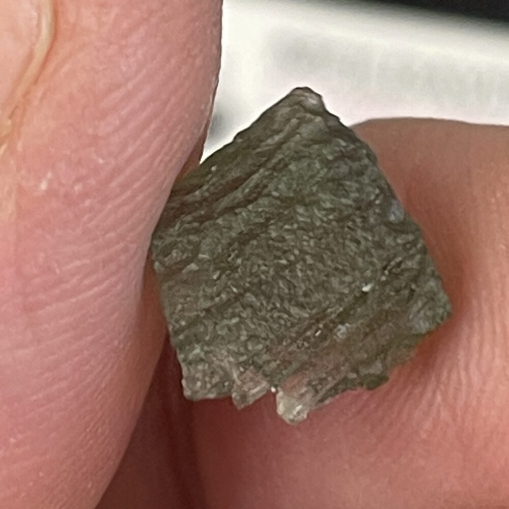 A close-up view of a hand holding a small, rough greenish-brown mineral crystal with a textured surface.