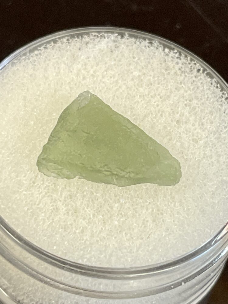 A pale green, rough triangular Moldavite From Chlum in CZ (.47g/2.50ct) is displayed on a white foam pad in a clear circular container.