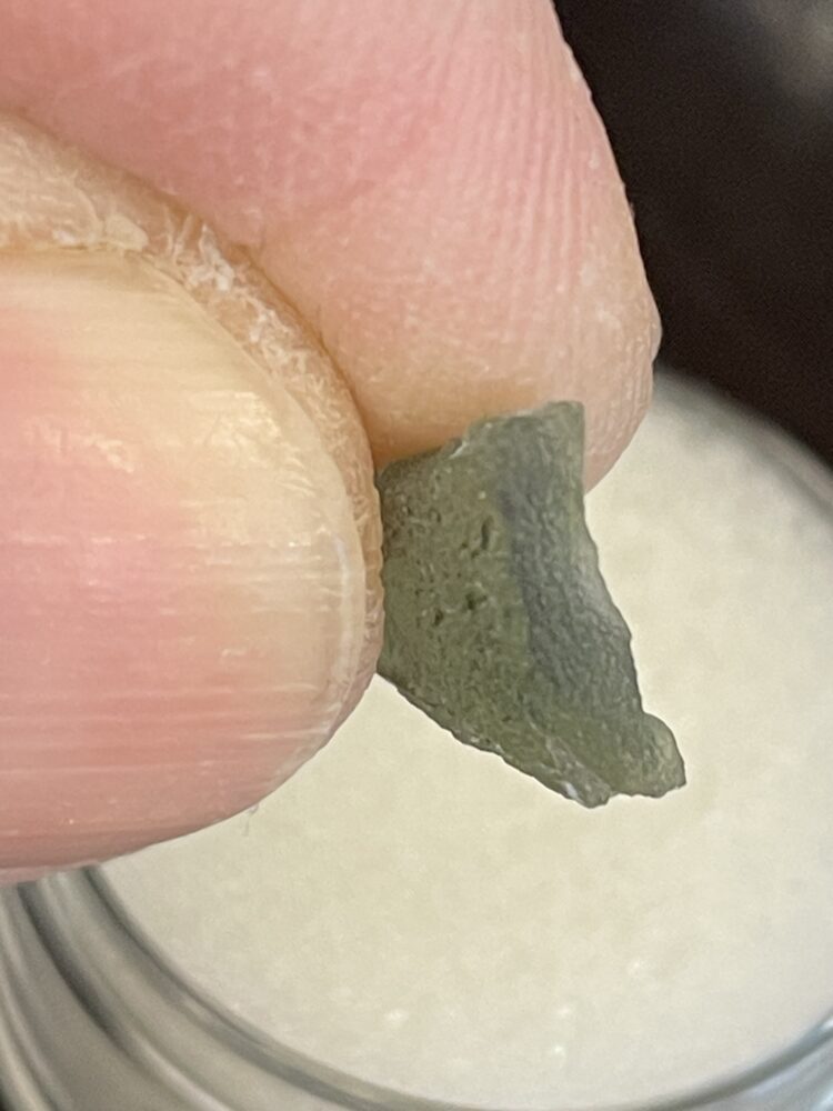 A close-up of fingers holding a .47g (2.50 ct) Moldavite From Chlum in CZ, highlighting its small size, irregular shape, and rough greenish-gray texture.