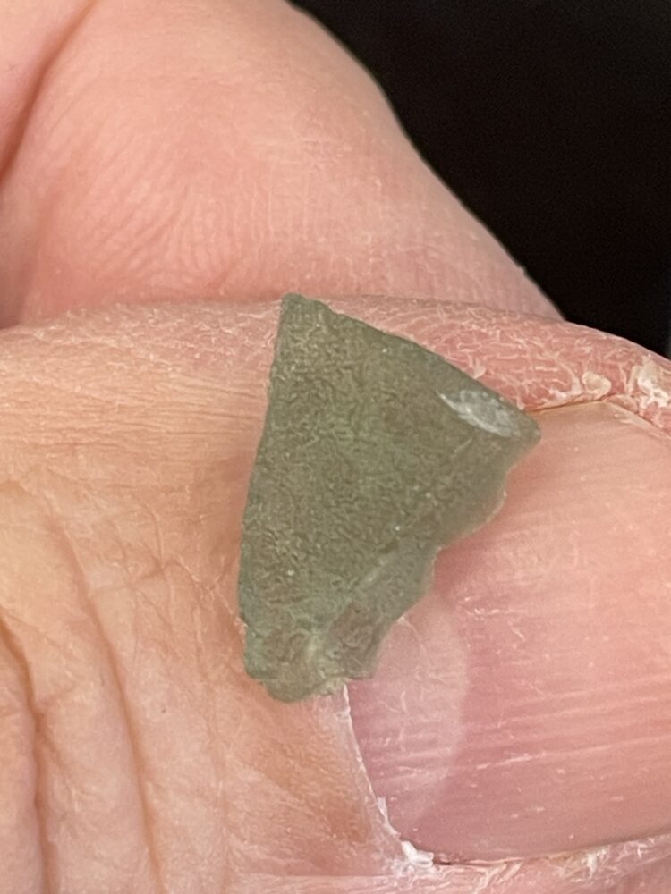 A close-up of fingers holding a small, greenish Moldavite From Chlum in CZ (.47g / 2.50 ct), showing its semi-translucent texture.