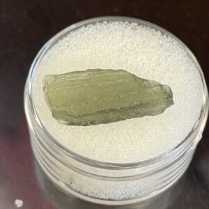 A greenish, rough Moldavite from Chlum in CZ is displayed in a transparent round container with a white foam base.