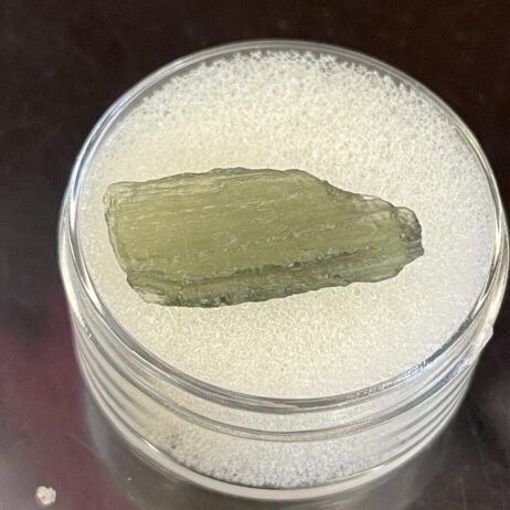 A greenish, rough Moldavite from Chlum in CZ is displayed in a transparent round container with a white foam base.