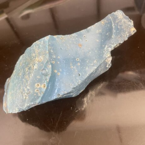 A rough, irregularly shaped blue stone with small holes and surface texture, resting on a reflective black surface.