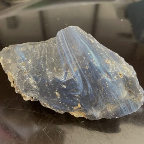 The Leland Blue Slag Glass, weighing 123g (4.3 oz), displays several blue shades, visible striations, and patches of dirt, its rough texture accentuated against a reflective dark surface.