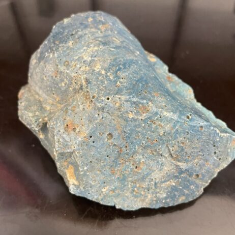 A rough, blueish rock with a textured surface and small holes sits on a reflective dark surface.
