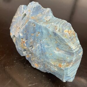 A rough, blue mineral specimen with small holes and brownish spots, displayed on a dark reflective surface.