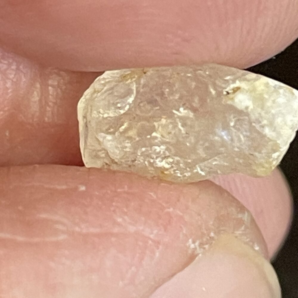 A close-up of fingers holding the Phenakite - Phenacite Crystal from Brazil 4.90 ct, highlighting its rough, translucent, and uneven yellowish surface.