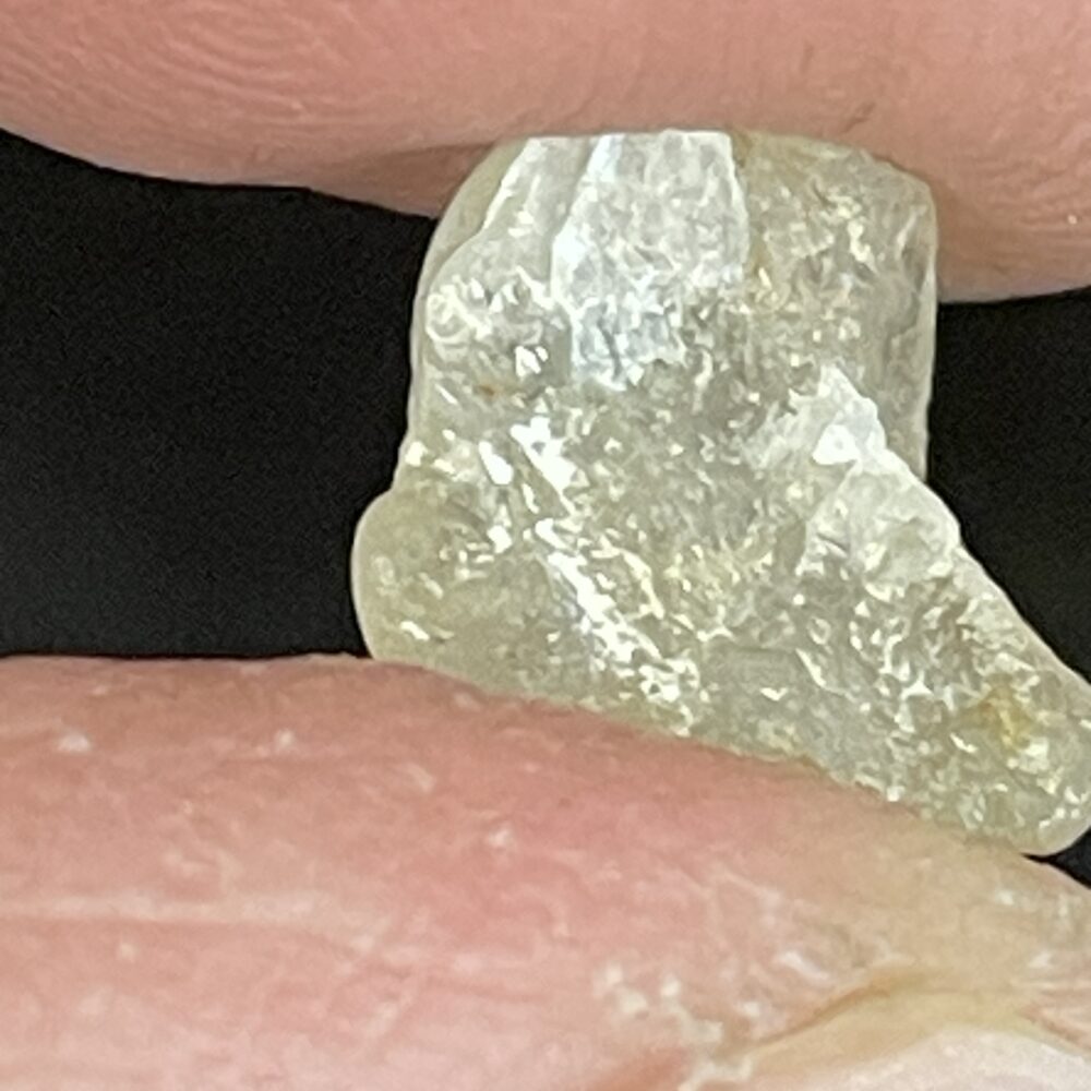 A rough, translucent gemstone or mineral is held between two fingers, showing uneven and jagged surfaces.
