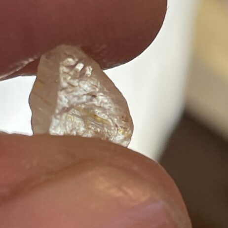 A close-up of two fingers holding a translucent, uncut gemstone or mineral fragment against a blurred background.