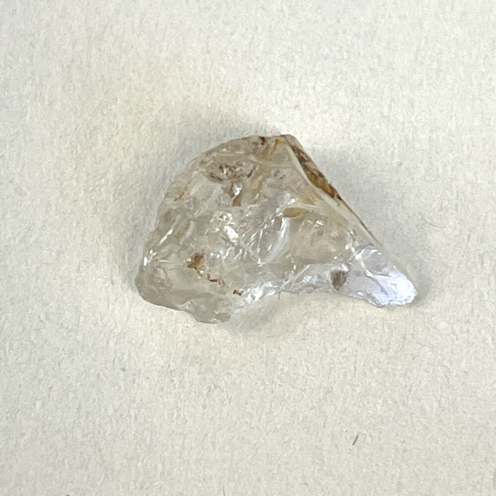A small, irregularly-shaped clear crystal with some brownish inclusions resting on a plain white surface.
