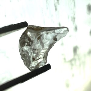 A close-up of metal tweezers holding a rough, uncut, translucent gemstone against a light background.