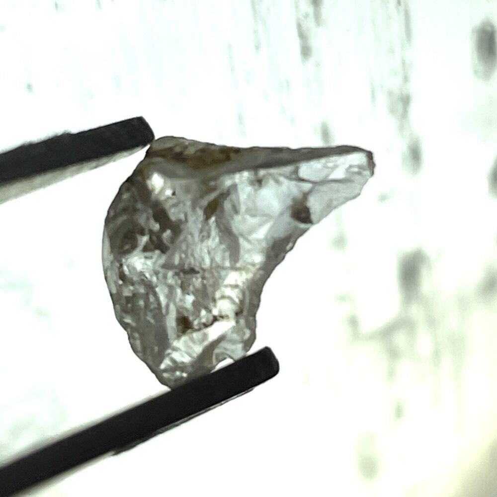A close-up of metal tweezers holding a rough, uncut, translucent gemstone against a light background.