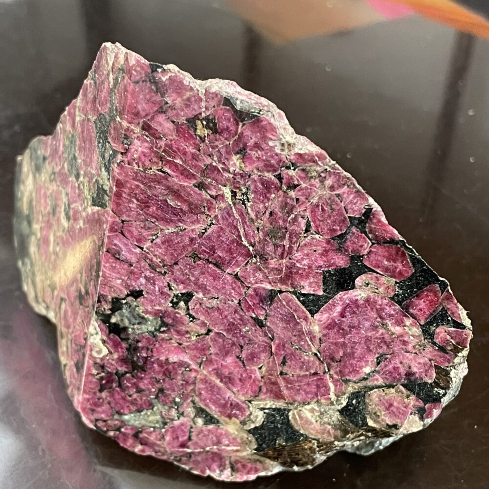 A rough mineral specimen featuring clusters of dark pink crystals with black and greenish matrix, displayed on a reflective surface.