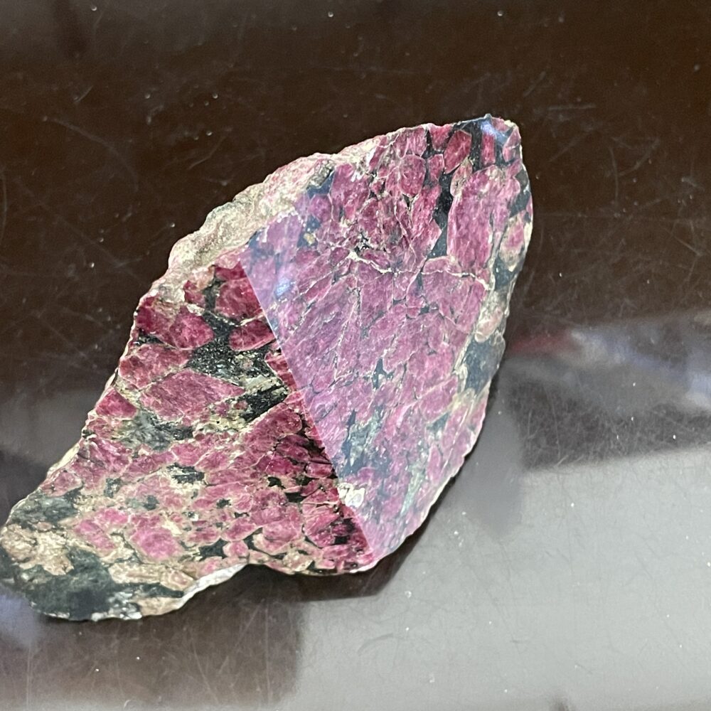 A rough mineral sample with a polished triangular section, showing a surface of pinkish-red and black mottled patterns, placed on a reflective dark background.