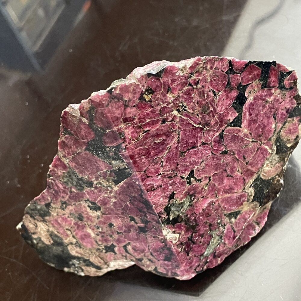 A rough, unpolished rock featuring clusters of deep red and pink mineral crystals with some black and greenish areas, displayed on a dark surface.