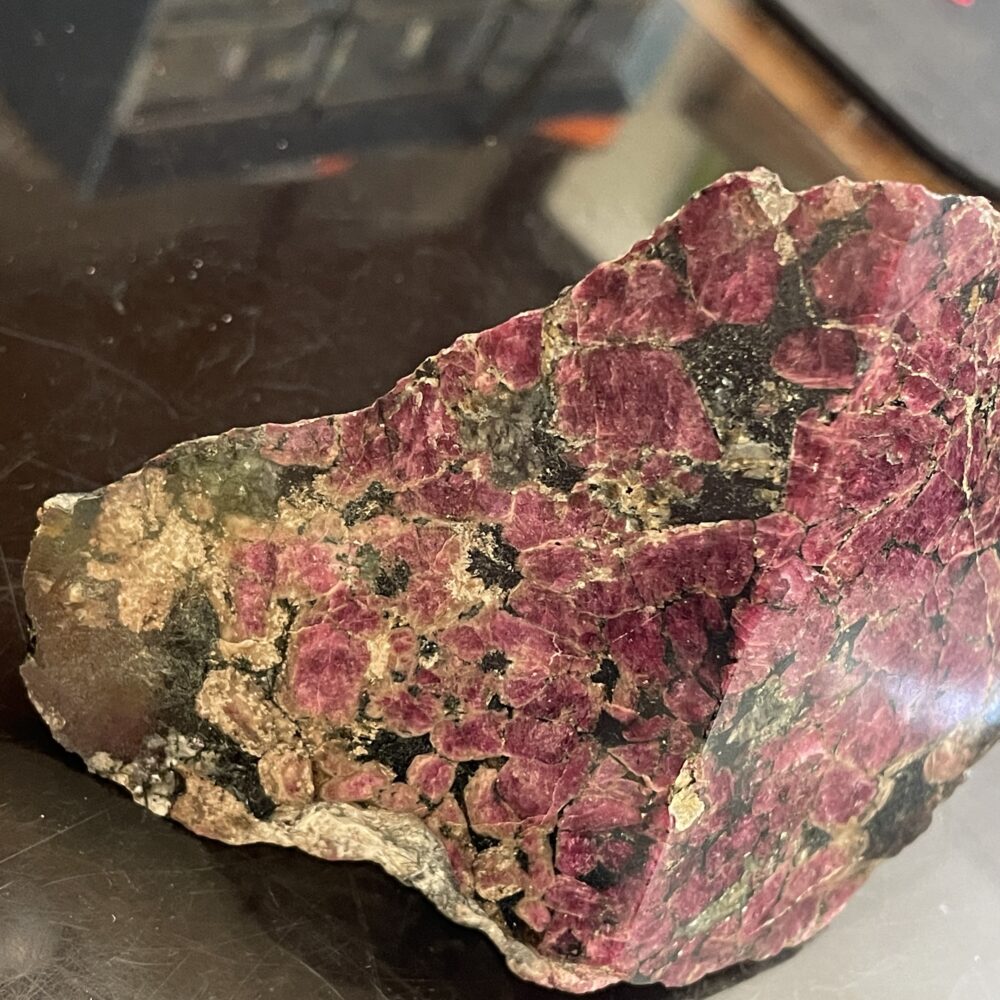 A rough, unpolished rock with dark green and purple-red mineral crystals on a reflective black surface.
