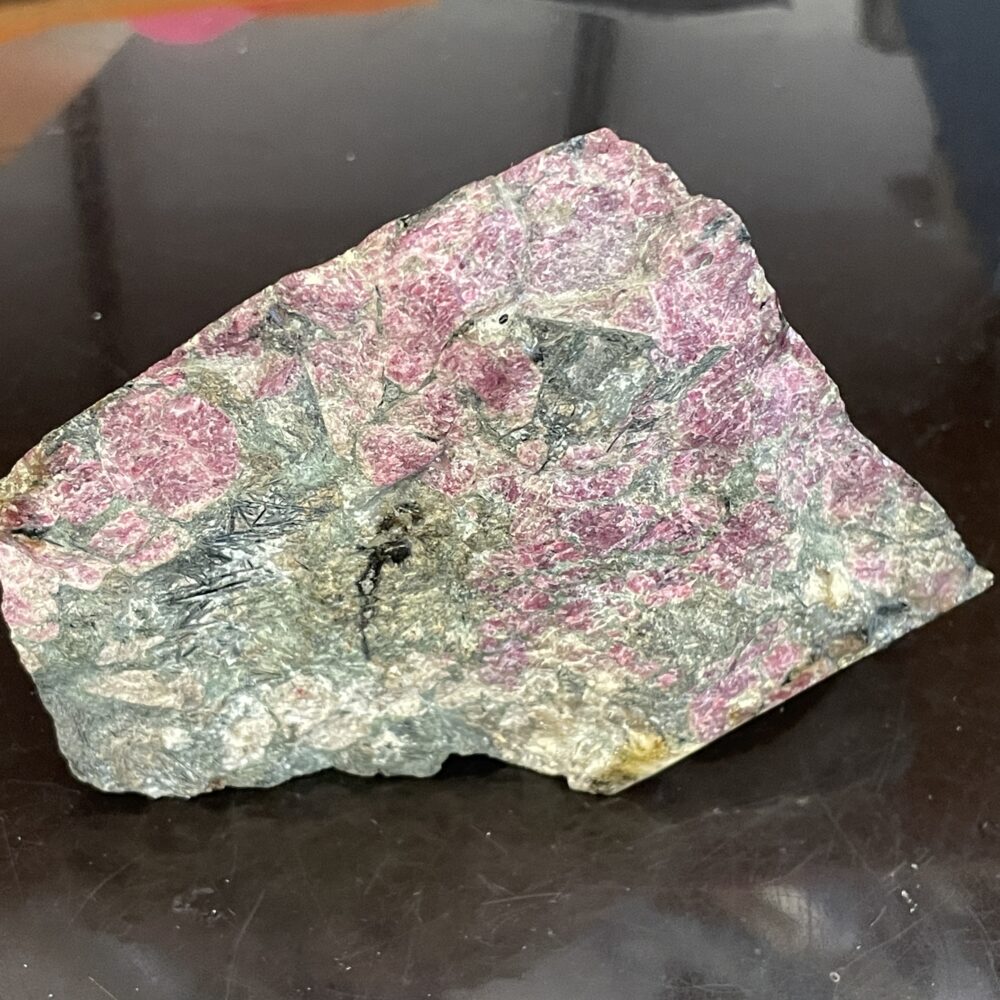 A rough, unpolished rock with prominent pink and grey mineral patches resting on a dark reflective surface.